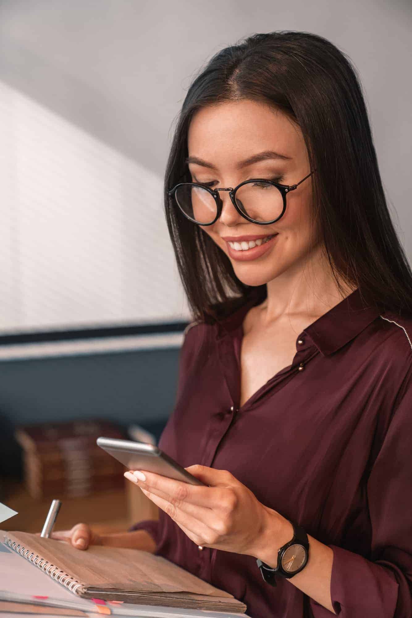 Cheerful female executive in glasses reading her messages on mobile phone with note book in hands